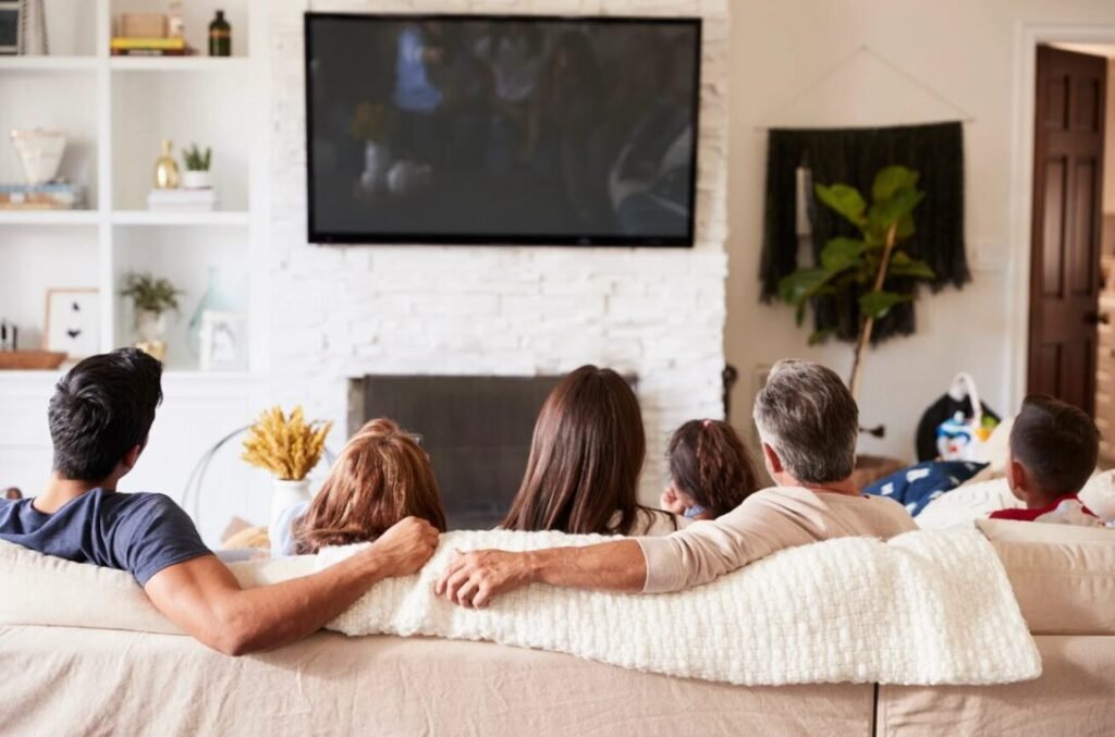 A family of six, including children and adults, sitting closely together on a couch watching TV in a cozy living room.