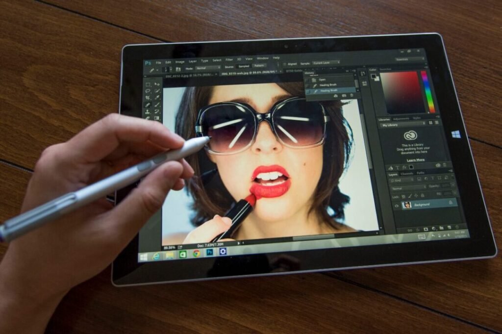 Hand editing a photo of a woman applying lipstick on a digital tablet using a stylus, with a photo editing software interface visible.