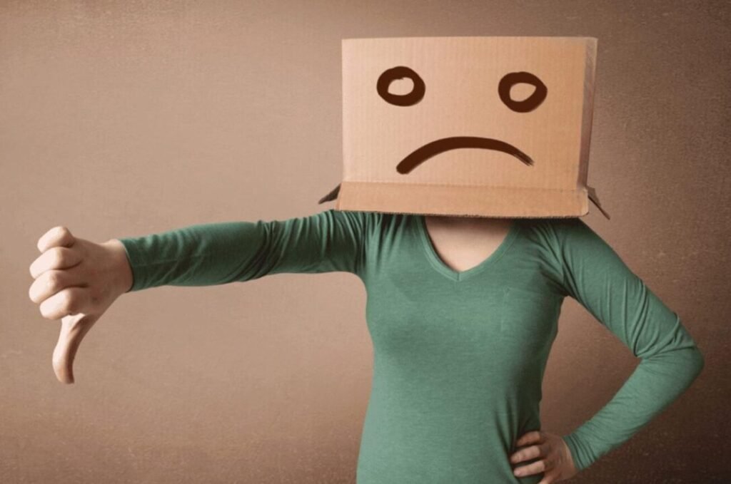 Person with cardboard box on their head with a sad face drawn on it, giving a thumbs-down gesture.