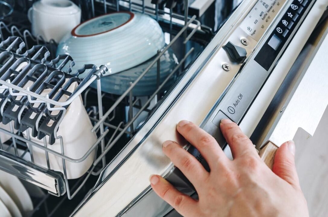 Person's hand closing a dishwasher full of clean dishes, showing control panel with buttons.