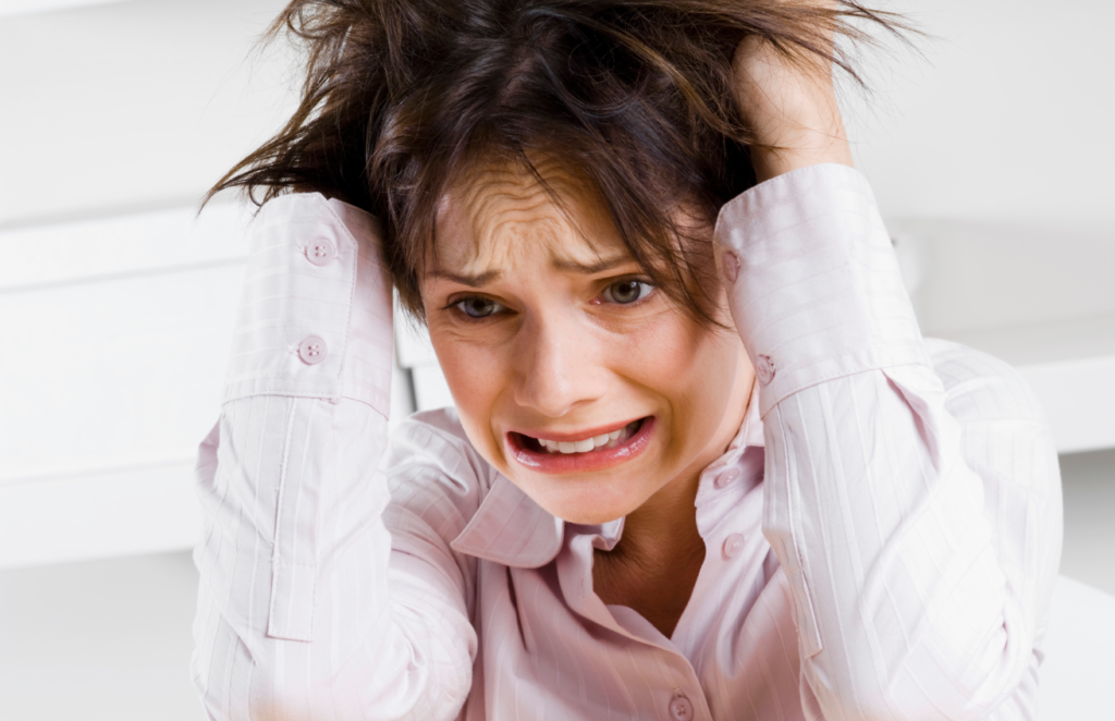 A distressed woman with both hands on her head, mouth open, expression showing panic or exasperation, wearing a light pink vertically striped shirt.