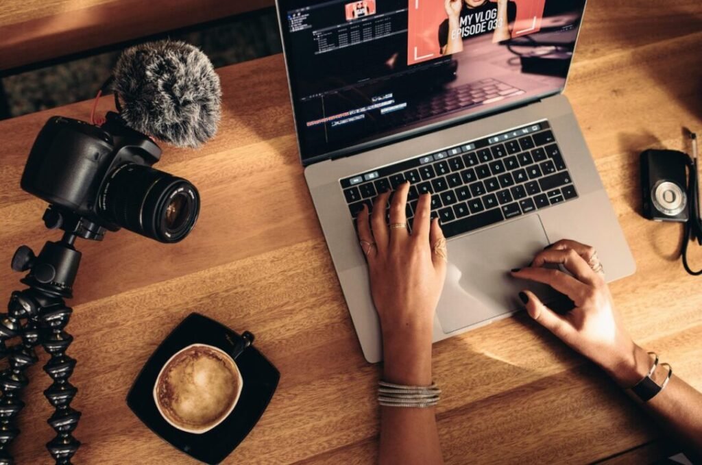 Overhead view of hands editing a vlog on a laptop, with a DSLR camera and coffee cup on a wooden table.