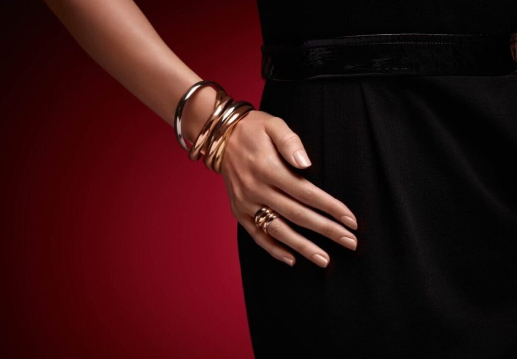 Woman's hand adorned with multiple golden bracelets and rings resting elegantly on her hip, wearing a black dress with a shiny belt, against a red background.