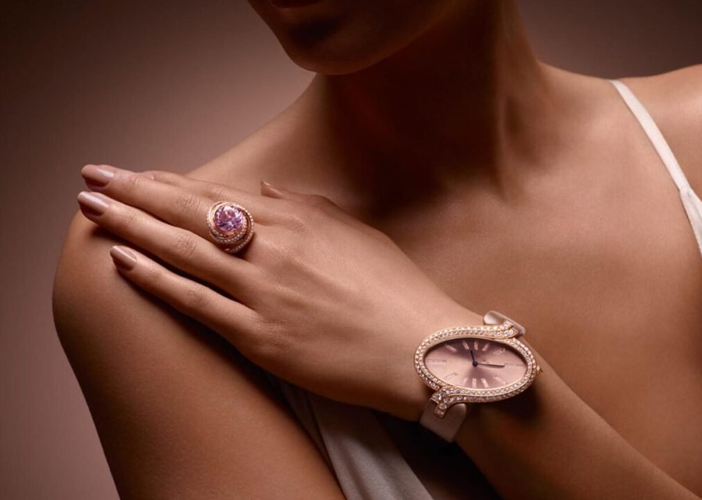 Woman showcasing elegant jewelry, wearing a large pink gemstone ring and a matching wristwatch with diamonds on a muted background.