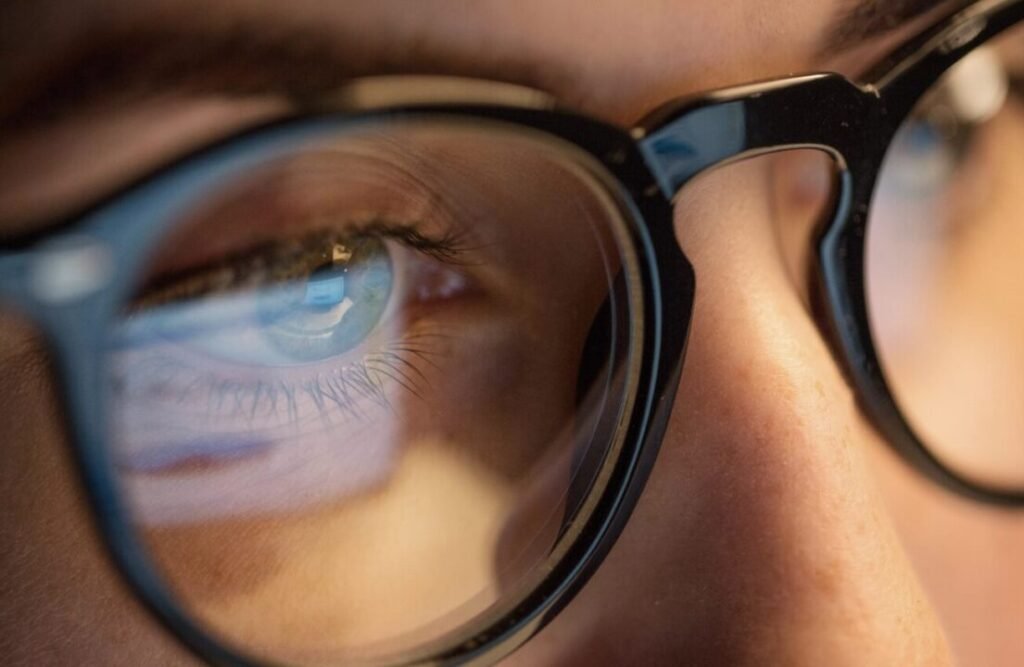 Intimate eye reflection in black-framed glasses, hinting at digital screen.