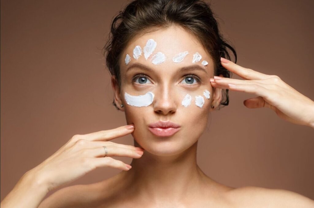 Woman with various patches of facial cream on her face, looking directly at the camera with a subtle expression, against a neutral brown background.