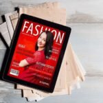 Fashion magazine cover on tablet, resting on newspapers, featuring smiling woman in red dress.