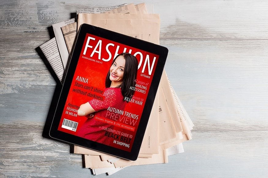 Fashion magazine cover on tablet, resting on newspapers, featuring smiling woman in red dress.