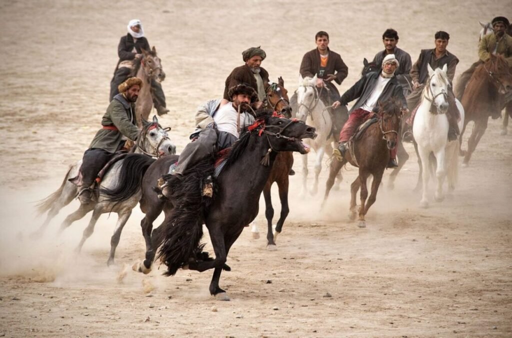 A group of men on horseback riding fast on a dusty ground, with some of the horses rearing and some riders leaning into the run.