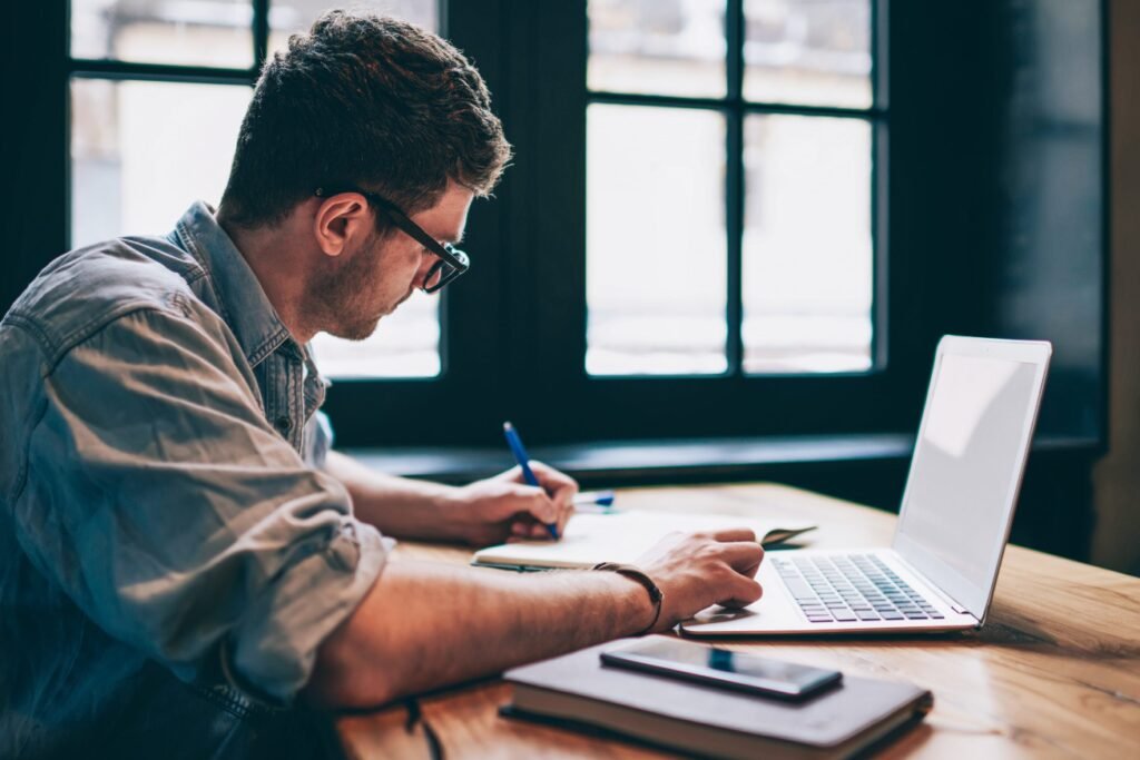 Professional man writing at wooden table with laptop and smartphone in bright office space.