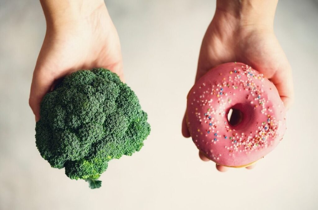 Two hands holding a broccoli head in one hand and a pink frosted donut with sprinkles in the other against a neutral background.