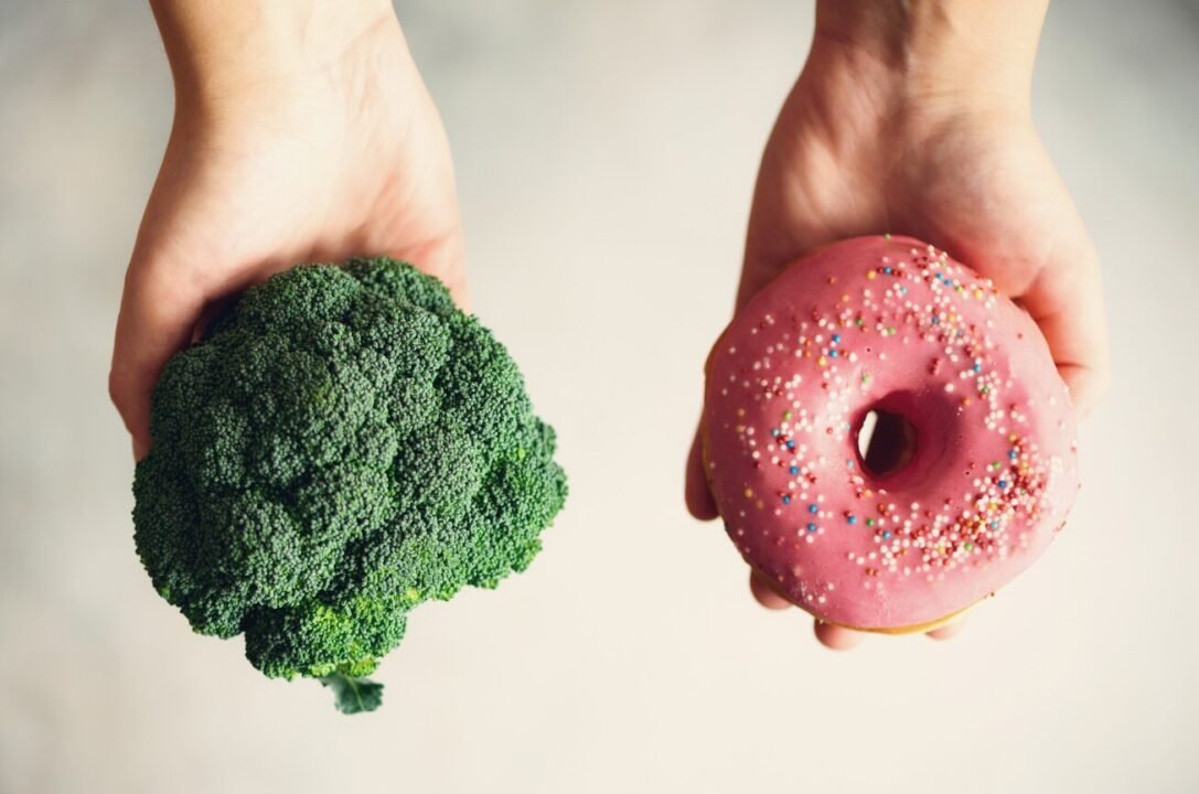 Healthy broccoli vs indulgent donut: food choice in hands on neutral background.