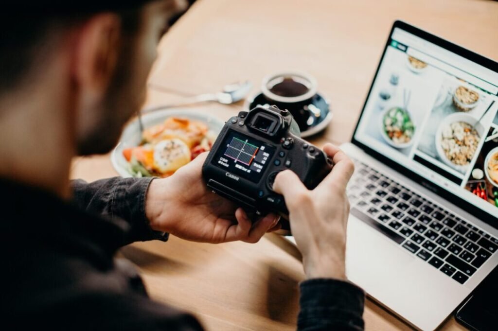 Person holding a DSLR camera, reviewing settings, with a laptop displaying food images and a plate of food on the table.