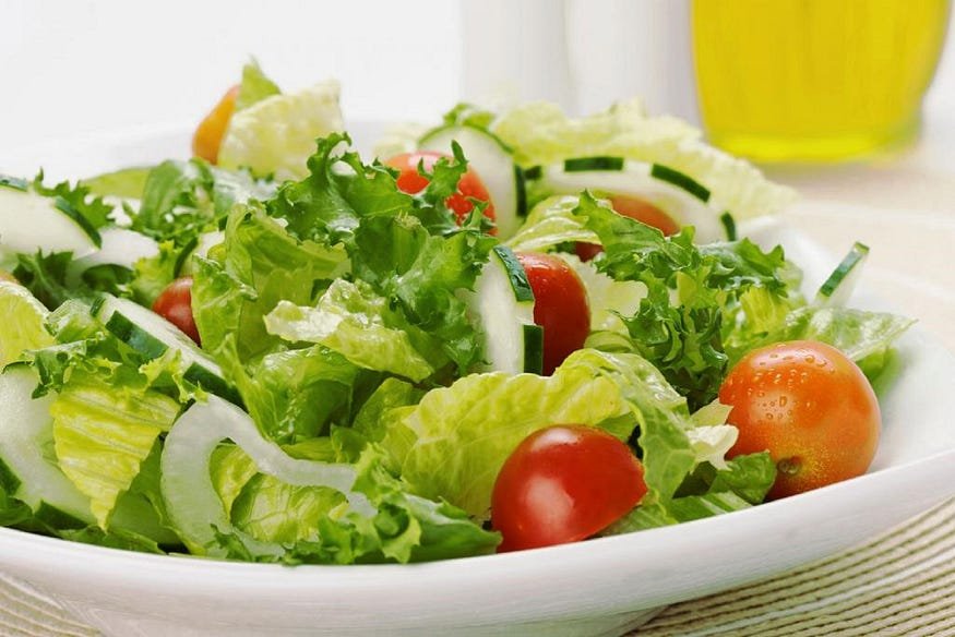 A fresh garden salad with lettuce, cherry tomatoes, cucumber slices, and chopped chives in a white bowl, with a bottle of dressing blurred in the background.