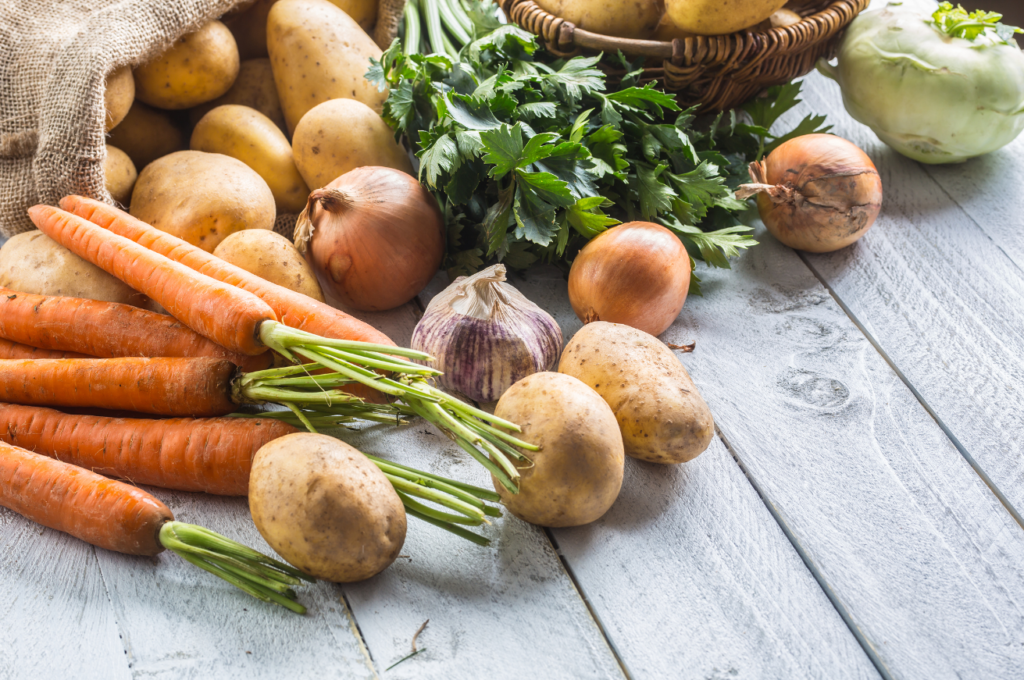 A variety of fresh vegetables including carrots, potatoes, onions, garlic, parsley, and kohlrabi on a rustic wooden surface.