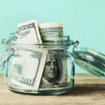 Glass jar with US $100 bills symbolizing savings on wooden surface against teal background.