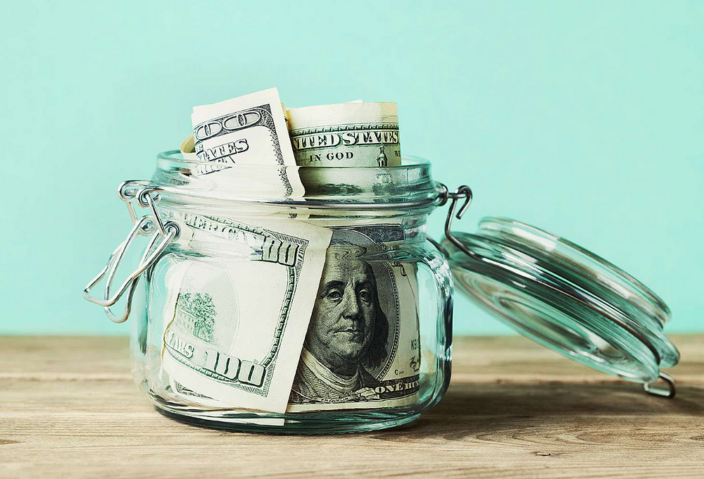 Glass jar with US $100 bills symbolizing savings on wooden surface against teal background.
