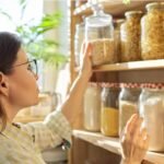 Woman selecting ingredients from organized kitchen pantry shelves with glass jars.