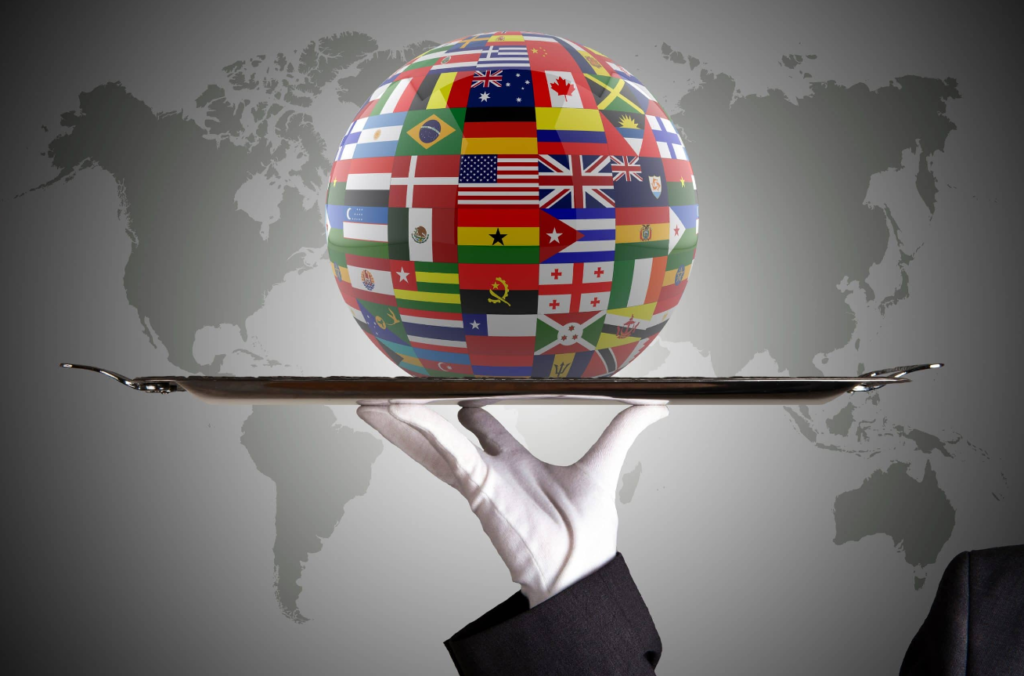 A person in formal attire holding a silver tray with a globe covered in various national flags in front of a world map silhouette on a gray background.