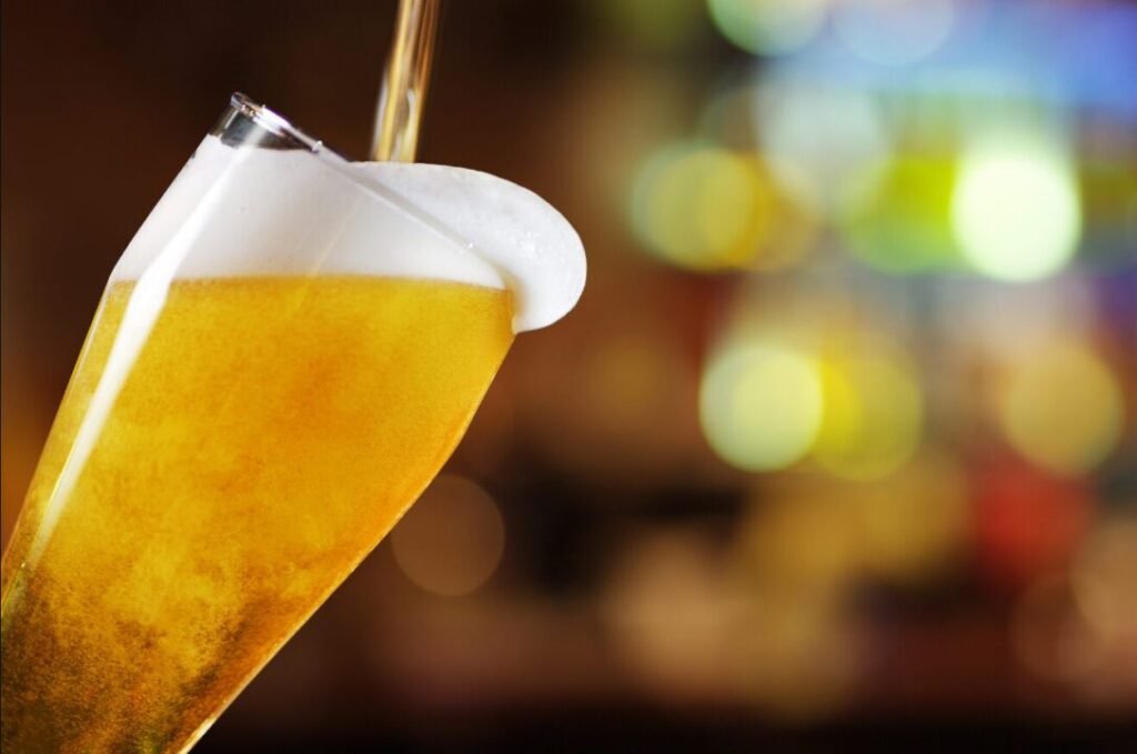 Golden beer pouring into tilted glass with frothy head, colorful bar background.