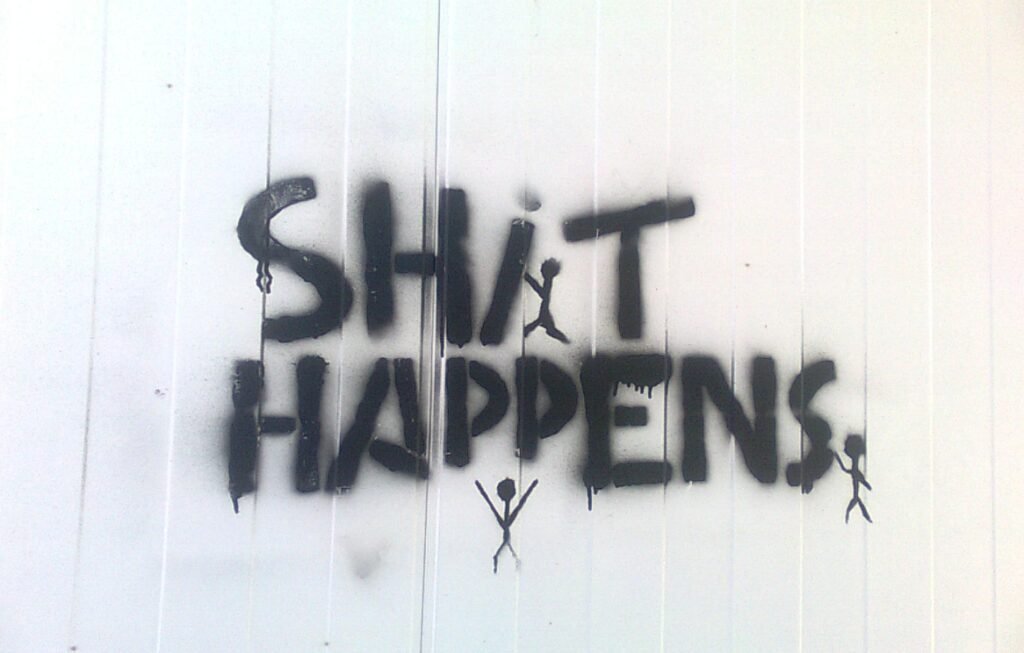 Black graffiti on a white corrugated surface reading "SHIT HAPPENS" with three stick figures underneath.