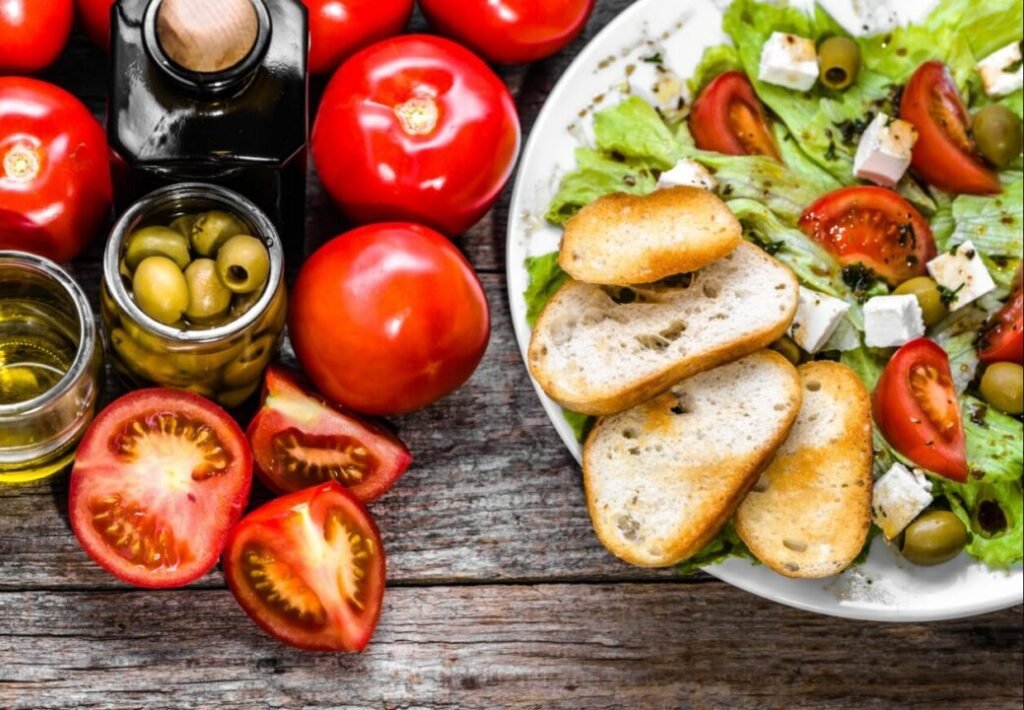 A plate of Greek salad with tomatoes, olives, feta cheese, and toasted bread, alongside whole tomatoes, a bottle of olive oil, and a jar of olives on a wooden table.