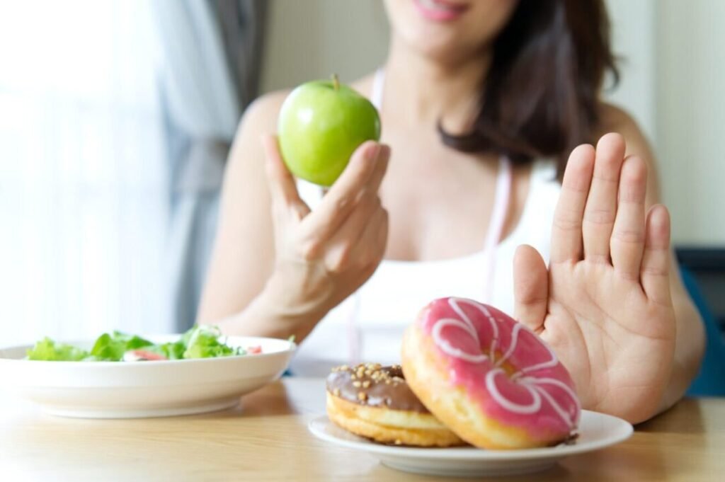 A woman holding a green apple in one hand and gesturing 'no' with the other hand, with a plate of colorful donuts in the foreground and a bowl of salad slightly out of focus in the background.