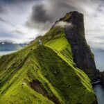 Majestic green cliff overlooking sea with white building, clouds, and mountains in background.