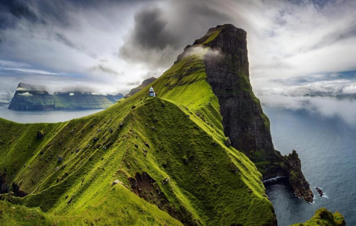 Majestic green cliff overlooking sea with white building, clouds, and mountains in background.