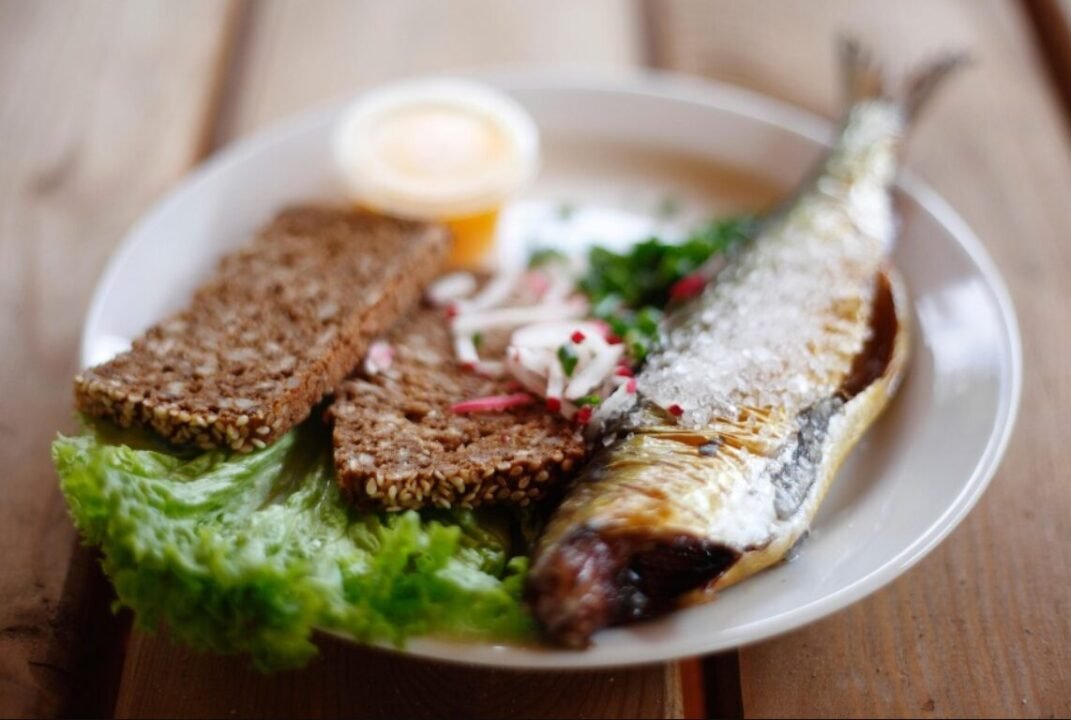 A plate of food featuring a grilled fish sprinkled with coarse salt beside slices of whole grain bread, a small side salad, and a cup of sauce.