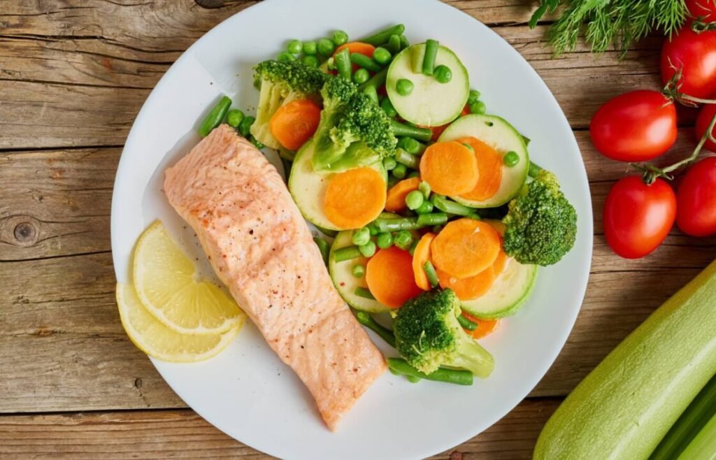 A grilled salmon fillet with a side of mixed vegetables including broccoli, green peas, carrots, and zucchini, garnished with lemon slices on a white plate, with fresh tomatoes and zucchini visible on the wooden table.