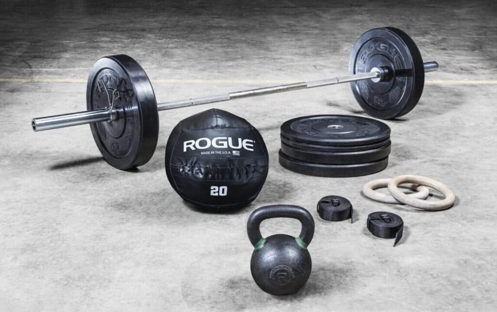 Various gym equipment on a concrete floor, including a barbell with weights, a black kettlebell, extra weight plates, a medicine ball labeled "ROGUE 20," and gymnastic rings.