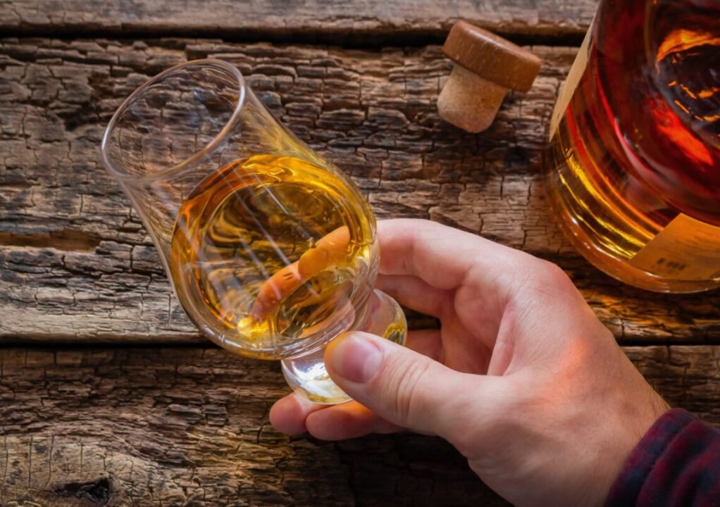 Hand holding a glass of whiskey on a rustic wooden table with a bottle next to it.