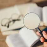 Hand holding magnifying glass over open book for close study and investigation.