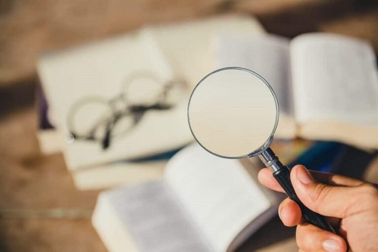 Hand holding magnifying glass over open book for close study and investigation.