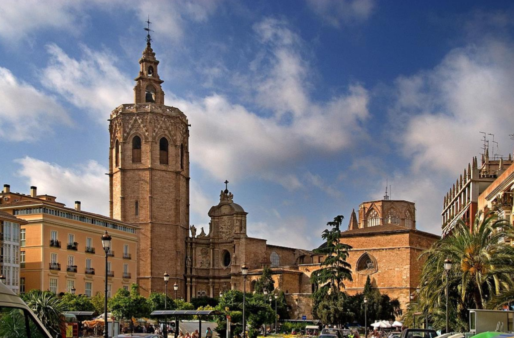 A view of a historic cathedral with a tall bell tower under a blue sky with clouds, flanked by a plaza with pedestrians and terrace seating, and lined with colorful buildings.