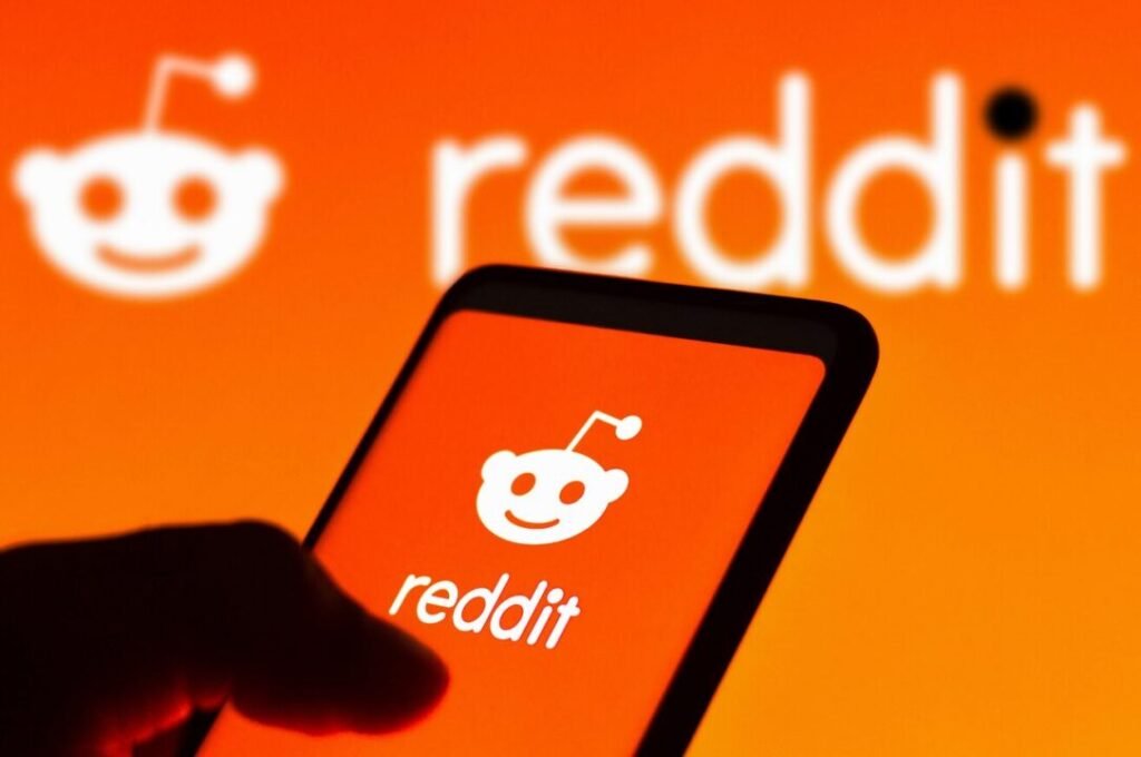 Person holding a smartphone displaying the Reddit logo, with a larger, out-of-focus Reddit logo in the background.