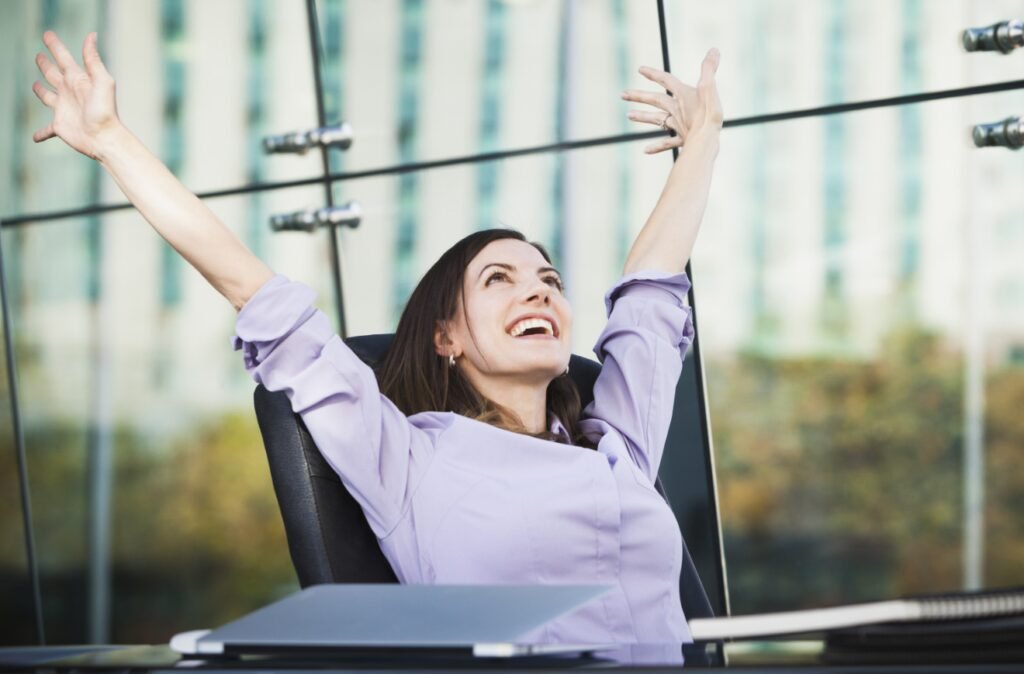 A joyful woman in a purple blouse stretching her arms up in the air while leaning back in an office chair, with a large glass window reflecting buildings in the background.