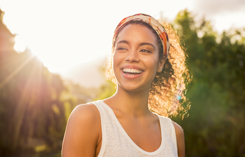 A joyful young woman wearing a headscarf with sun flare in the background.