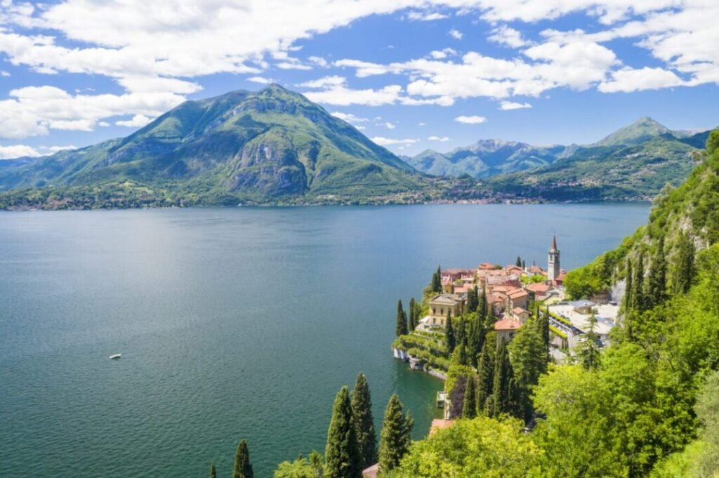 A panoramic view of a picturesque lakeside town with lush greenery, a prominent church steeple, surrounded by mountains under a partly cloudy sky.