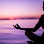 Tranquil meditation by the sea at sunset, person in lotus pose against colorful sky.
