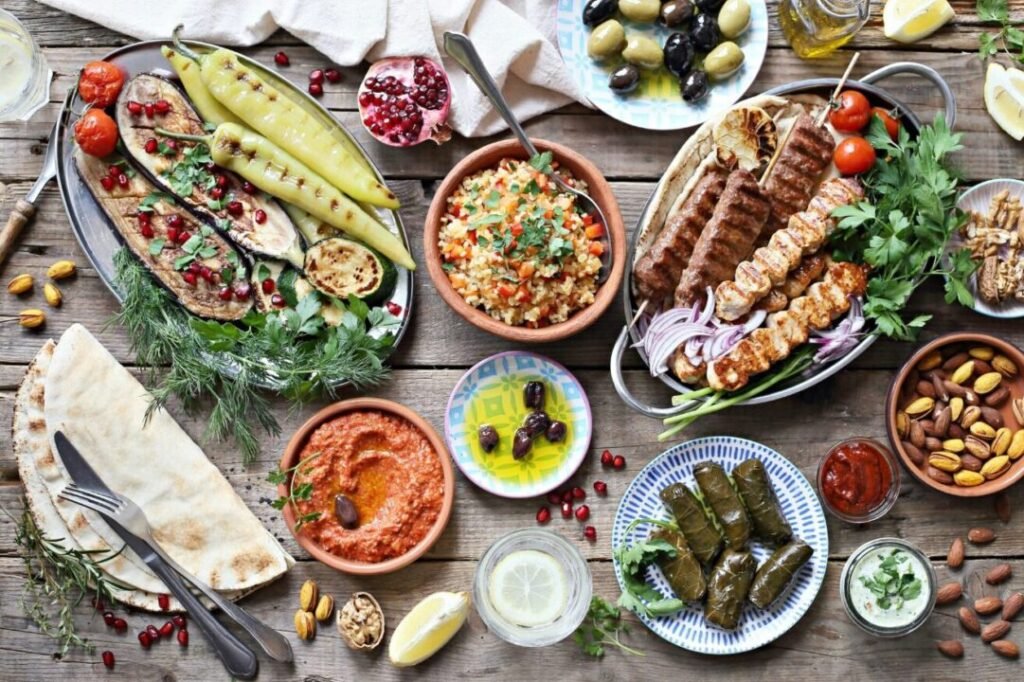 A variety of Mediterranean dishes spread on a wooden table, including grilled vegetables, kebabs, pilaf, dips, stuffed grape leaves, olives, nuts, and fresh herbs.