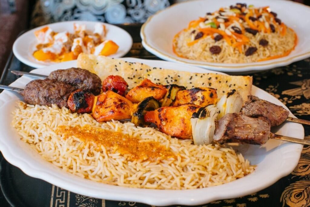 A plate of Middle Eastern cuisine with a variety of grilled kabobs, including beef, chicken, and vegetables, served over a bed of rice, accompanied by a flatbread and two additional plates with sides in the background.