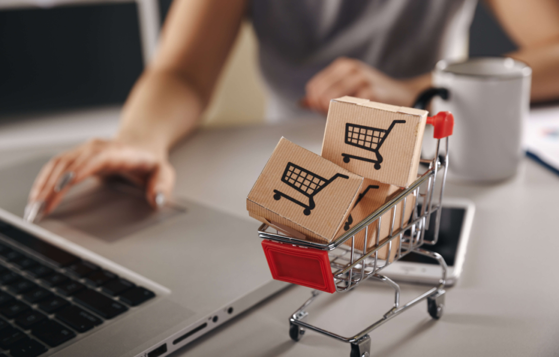 Mini shopping cart model on desk with laptop and mug symbolizing online shopping experience.