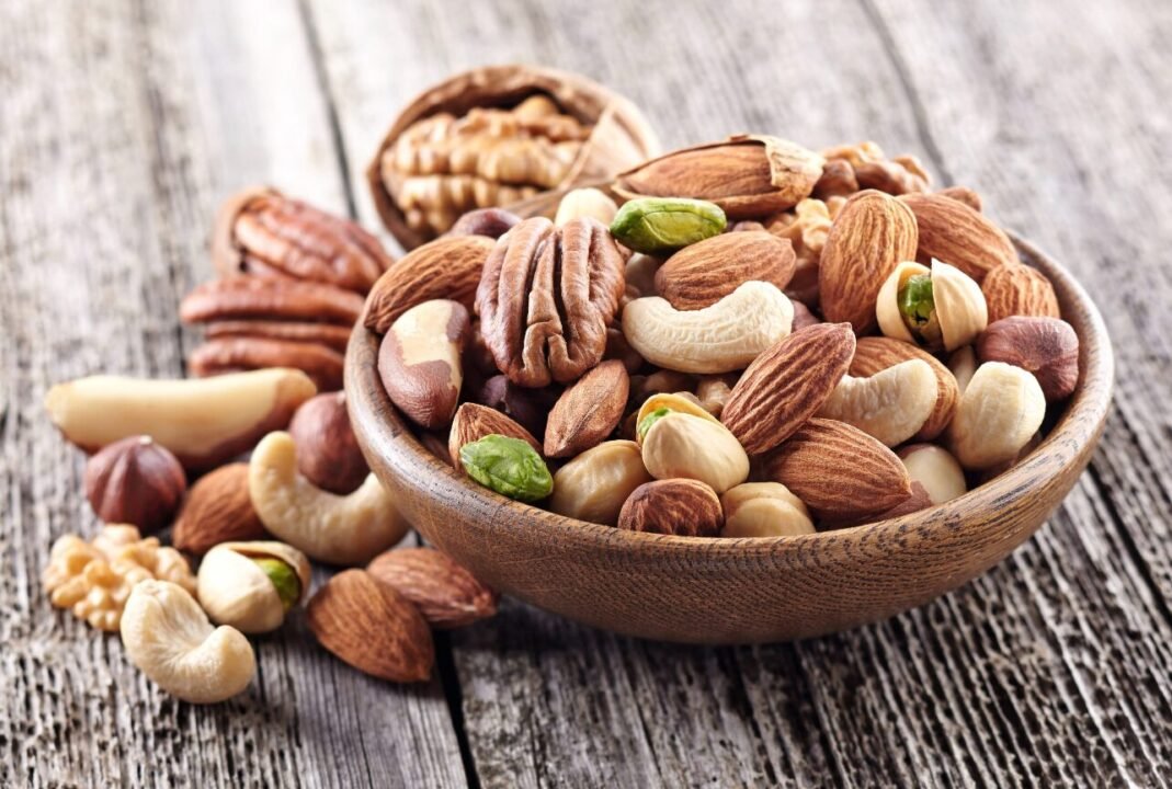 A wooden bowl filled with mixed nuts including almonds, cashews, hazelnuts, pecans, and pistachios, placed on a rustic wooden surface.