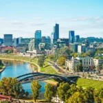 Tranquil river, modern city skyline with blend of contemporary and traditional architecture under clear sky.