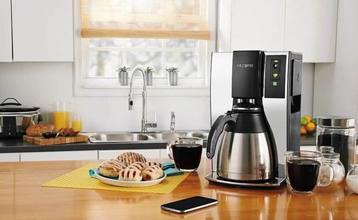 Modern kitchen counter with a coffee maker, glasses of coffee, a pitcher of orange juice, bread, and pastries on a wooden table, with a bright window and clean sink in the background.
