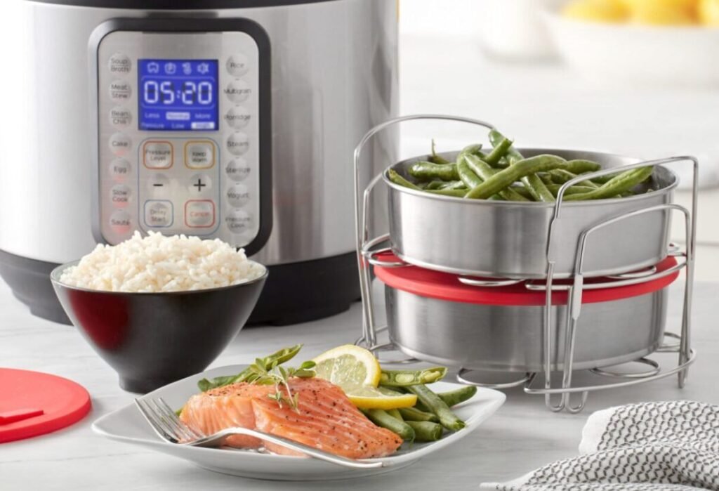 A modern kitchen setup featuring an electric pressure cooker with a digital display showing 05:20, surrounded by a plate of cooked salmon garnished with lemon and herbs, a bowl of rice, and stacked steaming pots with green beans.