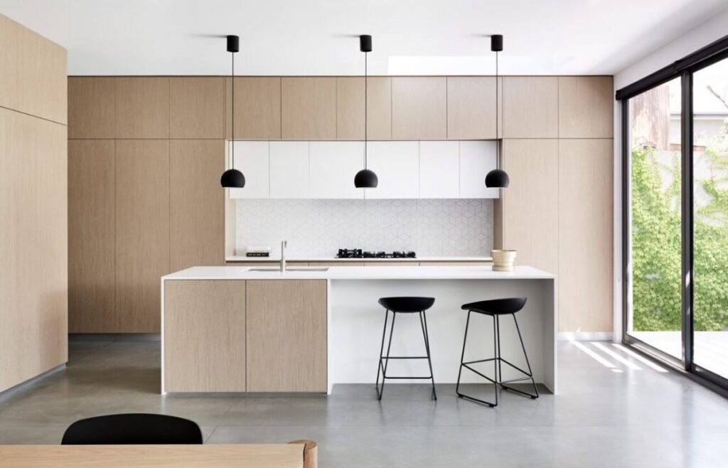 Modern kitchen interior featuring an island with bar stools, white and wooden cabinetry, suspended black lamps, and a large window with a view of greenery.