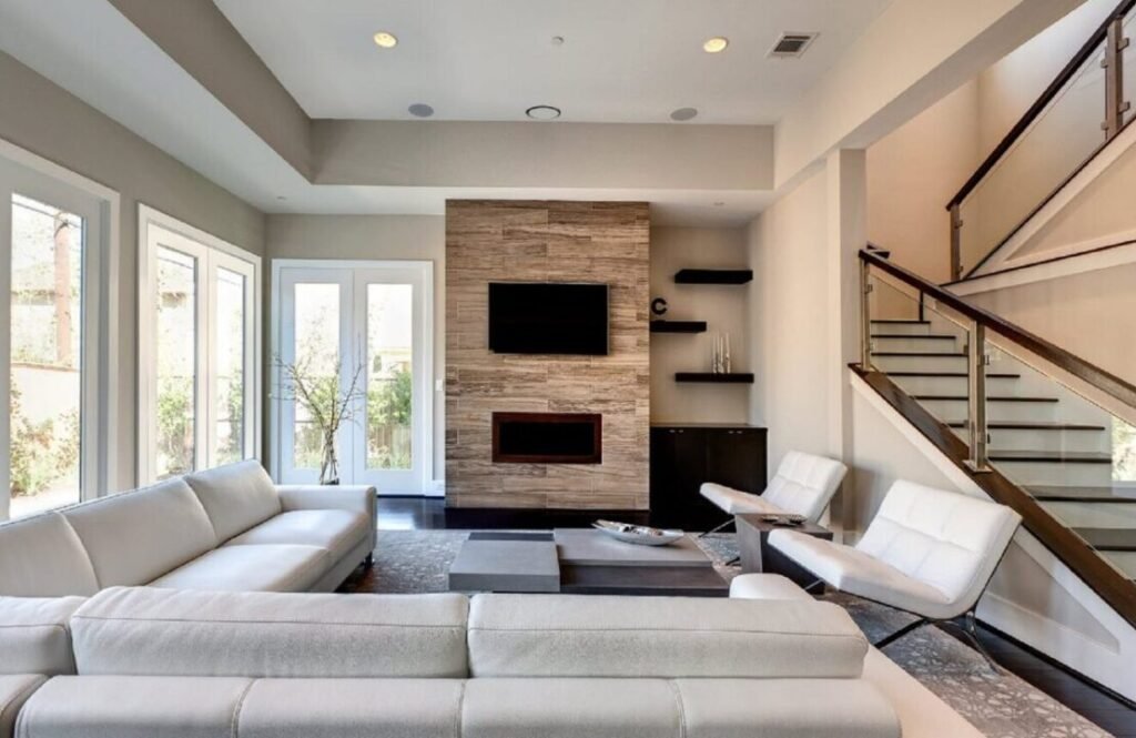 Modern living room with white sofas, a stone fireplace, wall-mounted TV, floating shelves, and a staircase with wood and glass railings.